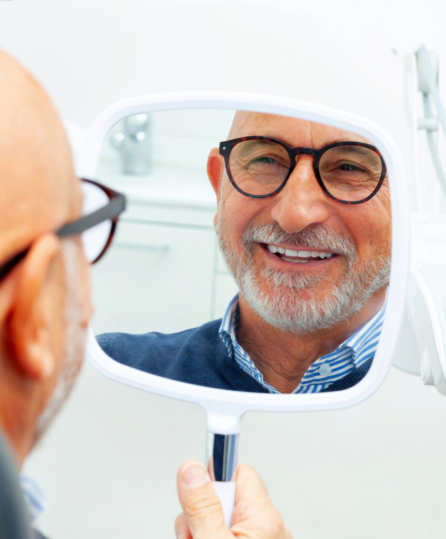 Happy senior man admiring his new smile in the small mirror at the dental clinic after a successful surgery