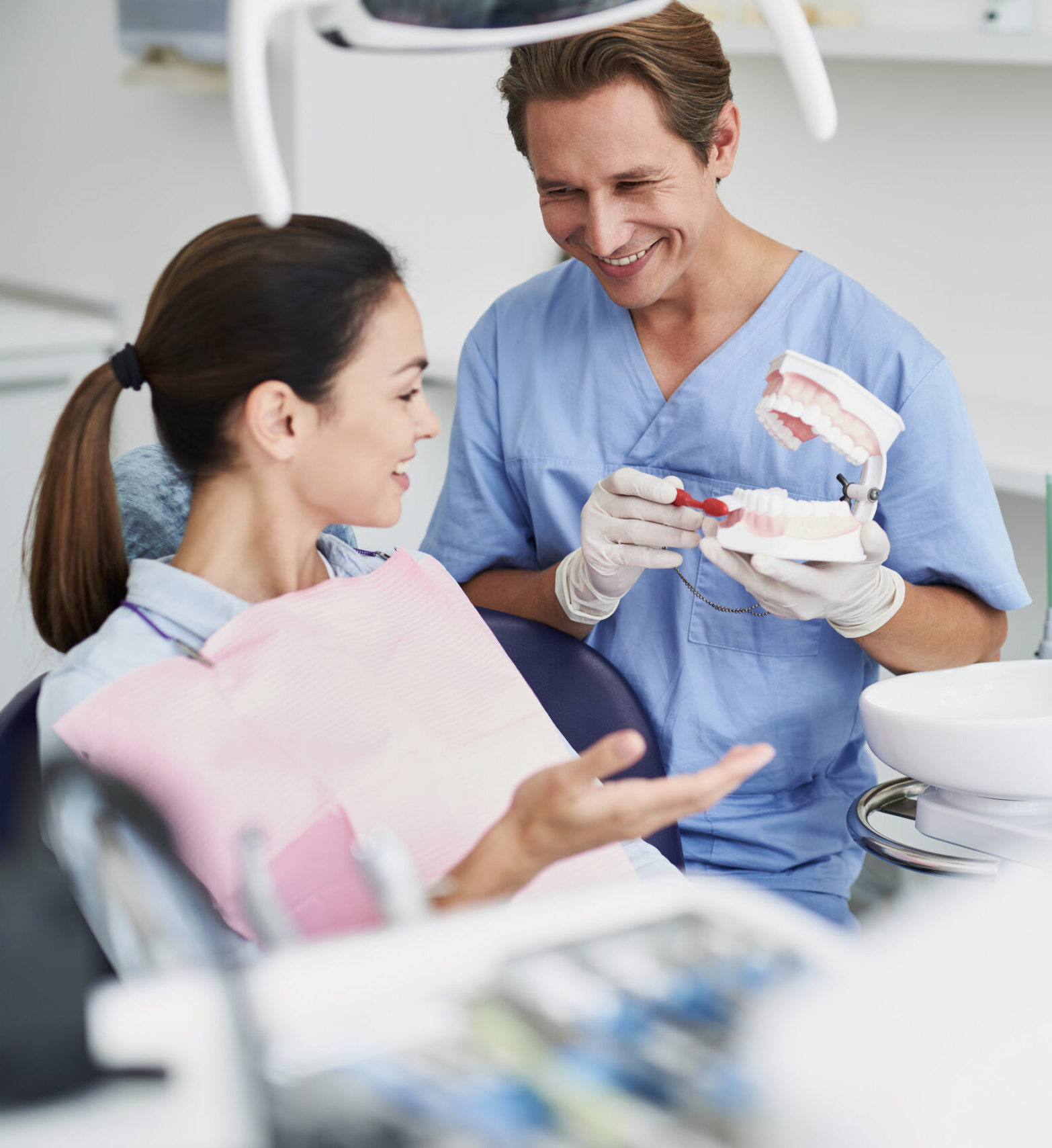 Consultation. Portrait of beautiful lady sitting in dental chair while stomatologist in sterile gloves holding false teeth and toothbrush