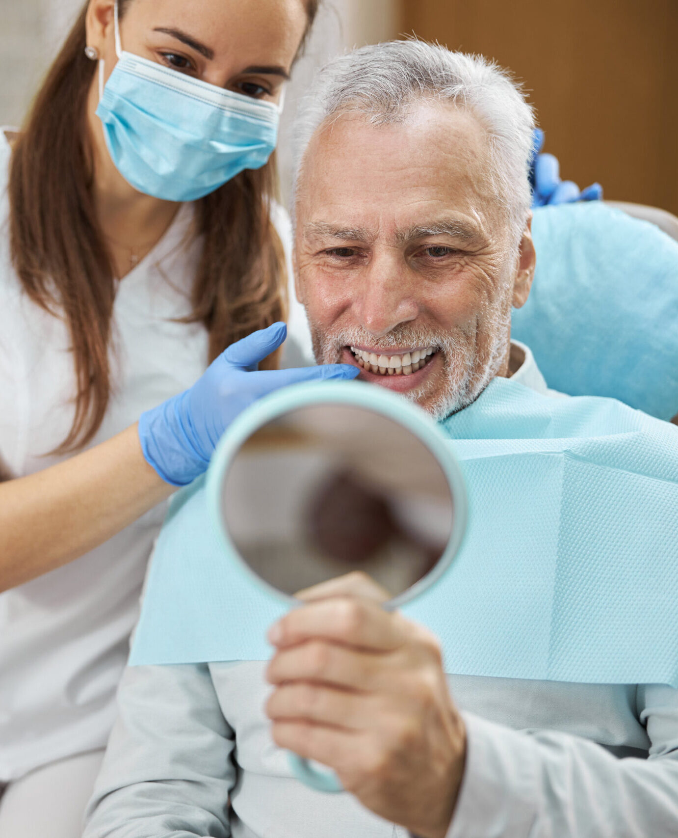 Elderly man and his young female dentist smiling while looking in the mirror in dental clinic