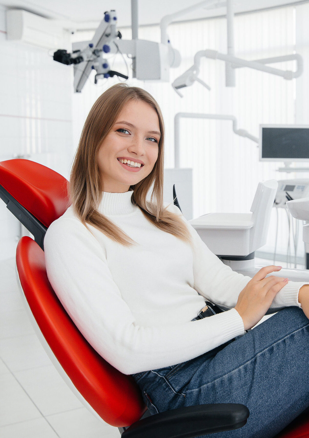 A beautiful young woman is sitting on a red dental chair in modern white dentistry and smiling. Dental prosthetics, treatment and teeth whitening.