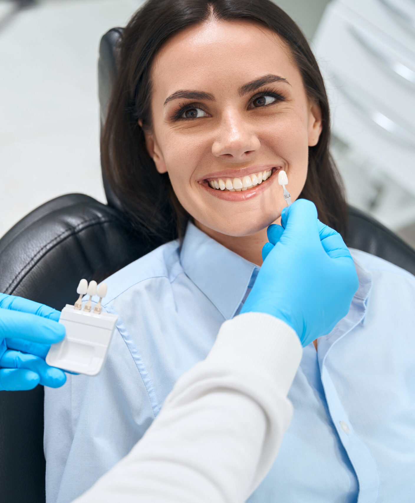 High-qualified dental technician choosing proper size and color of dental crown using some template, smiling woman client sitting in cozy orthodontic chair