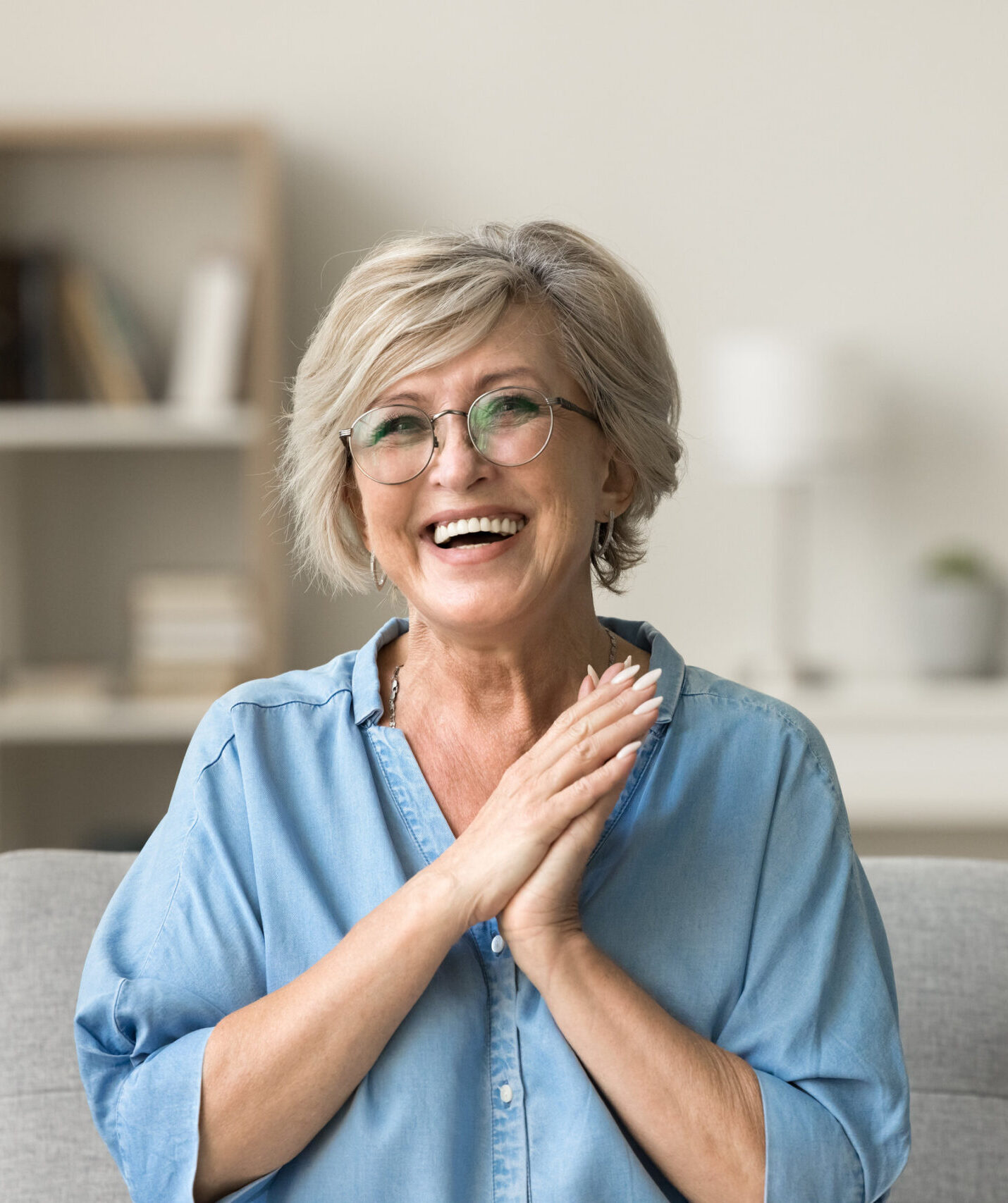 Cheerful pretty older woman in elegant glasses sitting on cozy home couch, smiling with perfect white teeth, laughing with hands at chest gesture, enjoying leisure, comfort, having fun